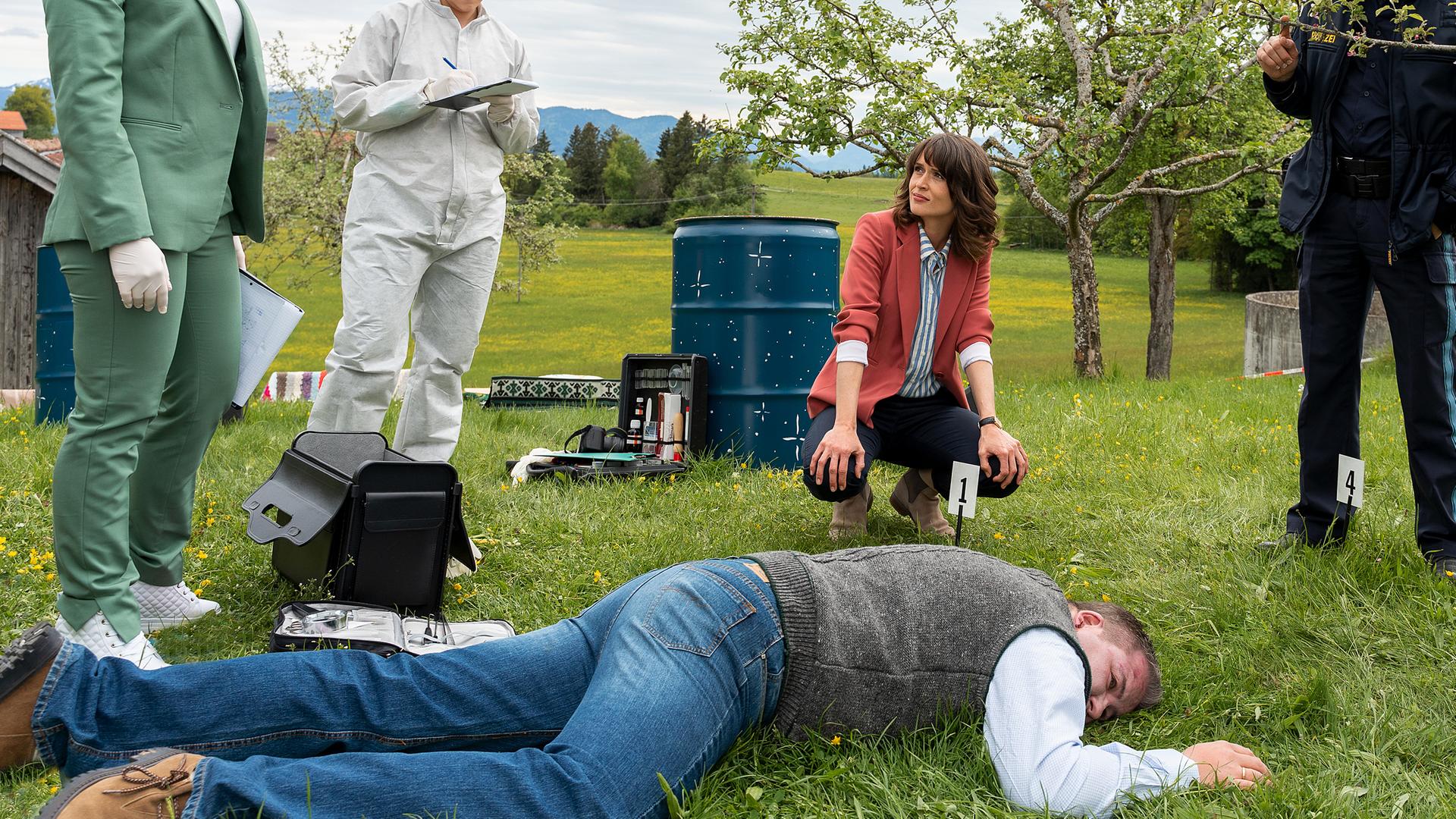 Ela Atay (Sevda Polat), die Spurensicherin (Julia Angeli), Kommissarin Andresen (Sophie Melbinger), Polizeihauptmeister Mohr (Max Müller) und Kommissar Stadler (Dieter Fischer) stehen auf einer Wiese im Halbkreis um das Opfer herum, das im Vordergrund des Bildes auf dem Bauch liegt. Im Hintergrund sind ein Regenfass sowie zahlreiche Bäume zu sehen.