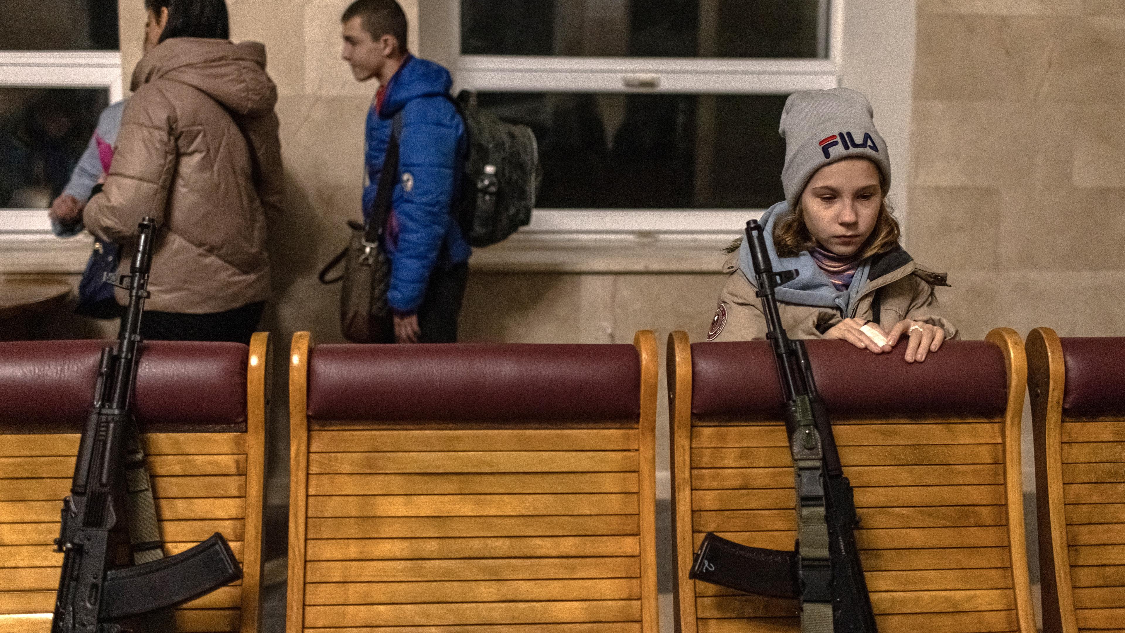 Ein Kind wartet auf einen Zug. Es lehnt dabei an eine niedrige Wand, an der zwei Gewehre stehen. 