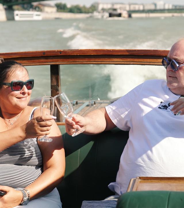 Waldi und Sonja sitzen bei sonnigem Wetter auf einem Boot und stoßen mit Sektgläsern an.