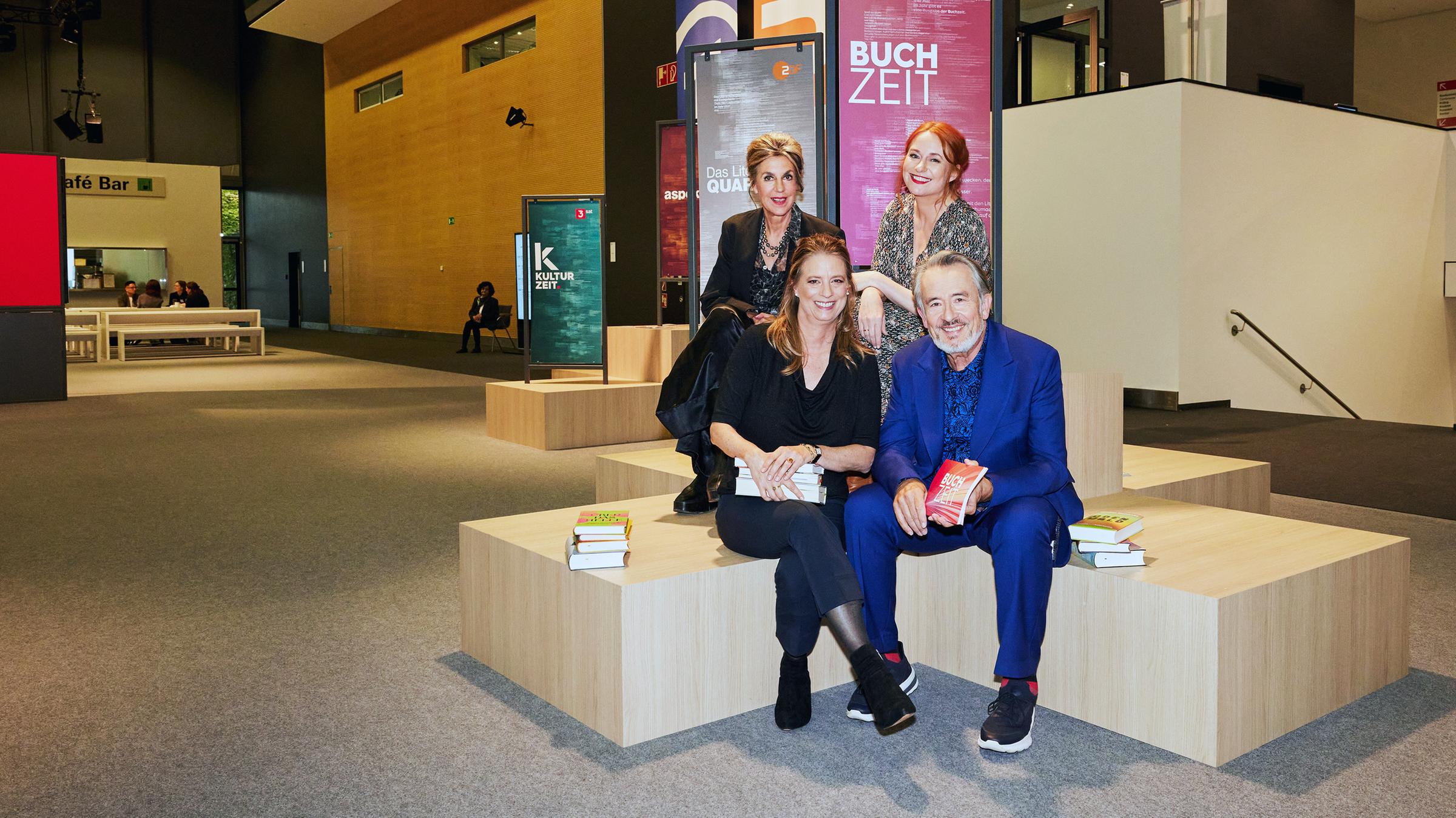 Moderator Gert Scobel und Sandra Kegel sitzen auf einem Podest. Dahinter, leicht erhöht sitzen Barbara Vinken und Katrin Schumacher ebenfalls auf einem Podest. Im Hintergrund sind Banner der Buchzeit, des Literarischen Quartetts und der Kulturzeit im Foyer der Frankfurter Buchmesse zu sehen.
