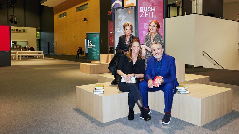 Moderator Gert Scobel und Sandra Kegel sitzen auf einem Podest. Dahinter, leicht erhöht sitzen Barbara Vinken und Katrin Schumacher ebenfalls auf einem Podest. Im Hintergrund sind Banner der Buchzeit, des Literarischen Quartetts und der Kulturzeit im Foyer der Frankfurter Buchmesse zu sehen.