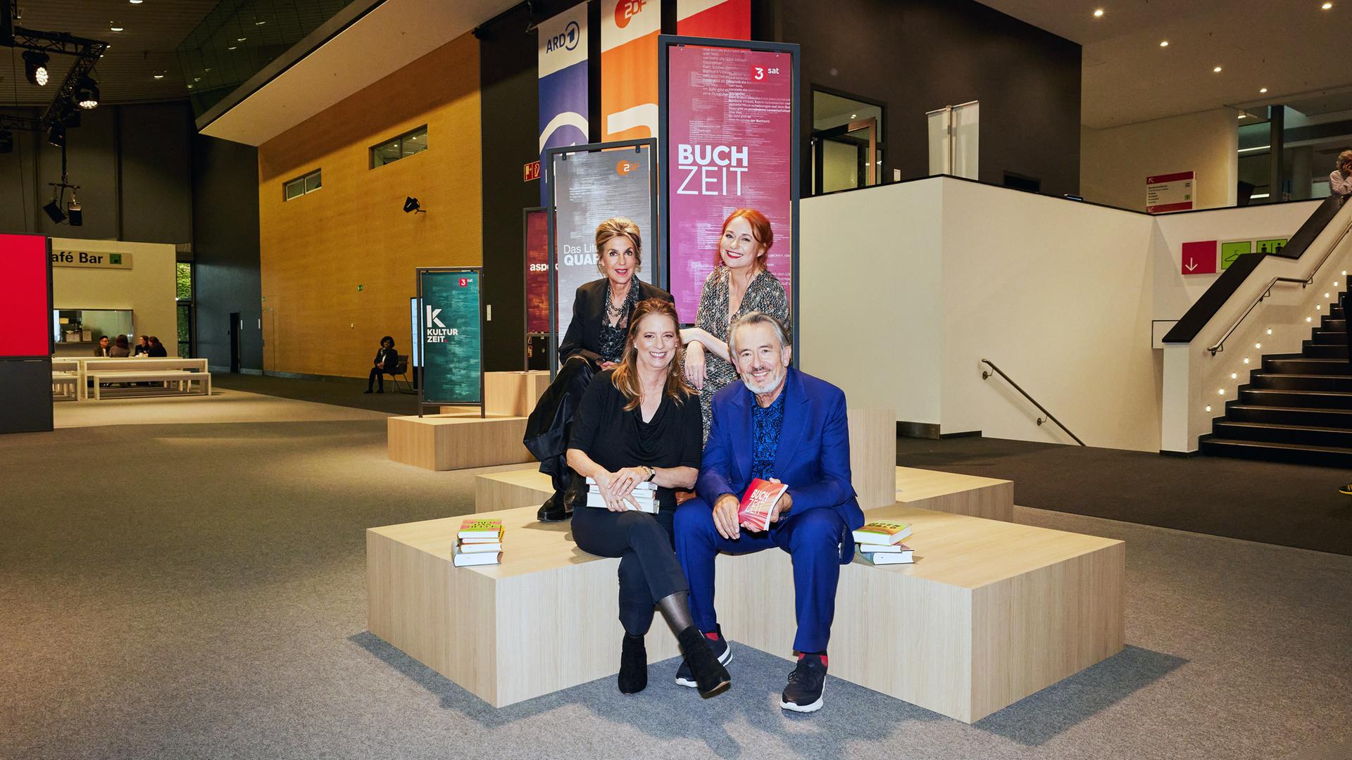 Moderator Gert Scobel und Sandra Kegel sitzen auf einem Podest. Barbara Vinken und Katrin Schumacher sitzen etwas erhöht hinter Scobel und Kegel ebenenfalls auf einem Podest. Im Hintergrund sind Banner der Buchzeit, des Literarischen Quartetts und der Kulturzeit im Foyer der Frankfurter Buchmesse zu sehen. 