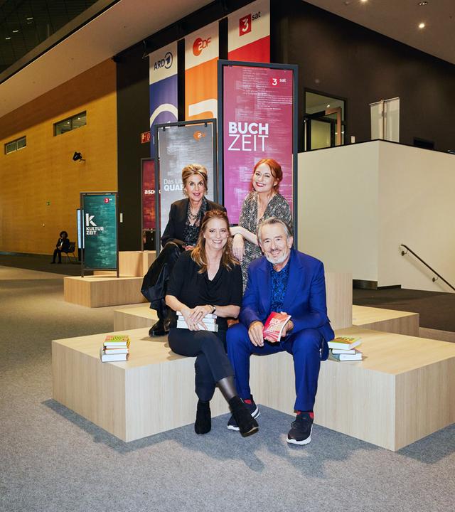 Moderator Gert Scobel und Sandra Kegel sitzen auf einem Podest. Barbara Vinken und Katrin Schumacher sitzen etwas erhöht hinter Scobel und Kegel ebenenfalls auf einem Podest. Im Hintergrund sind Banner der Buchzeit, des Literarischen Quartetts und der Kulturzeit im Foyer der Frankfurter Buchmesse zu sehen. 