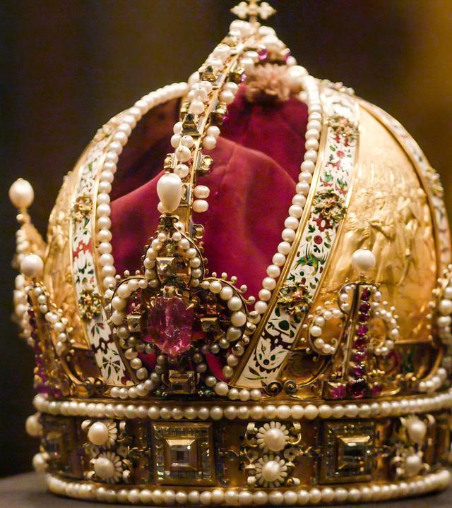 Das Bild zeigt eine prächtige Krone, die als Kaiserkrone bekannt ist. Sie ist aus goldfarbenem Material gefertigt und reich verziert mit Perlen, Edelsteinen und kunstvollen Verzierungen. In der Mitte der Krone befindet sich ein großer, rosa Edelstein, umgeben von weiteren glitzernden Steinen und schimmernden Perlen. Der obere Teil der Krone weist eine geschwungene Form auf und ist mit einer feinen Goldverzierung sowie zahlreichen kleinen Schmucksteinen dekoriert. Der Hintergrund ist dunkel und unscharf, wodurch die Krone im Vordergrund stark hervorsticht.   Diese Krone gehört zur Kaiserlichen Schatzkammer in der Wiener Hofburg und hat eine historische Bedeutung im Zusammenhang mit der Habsburgermonarchie.
