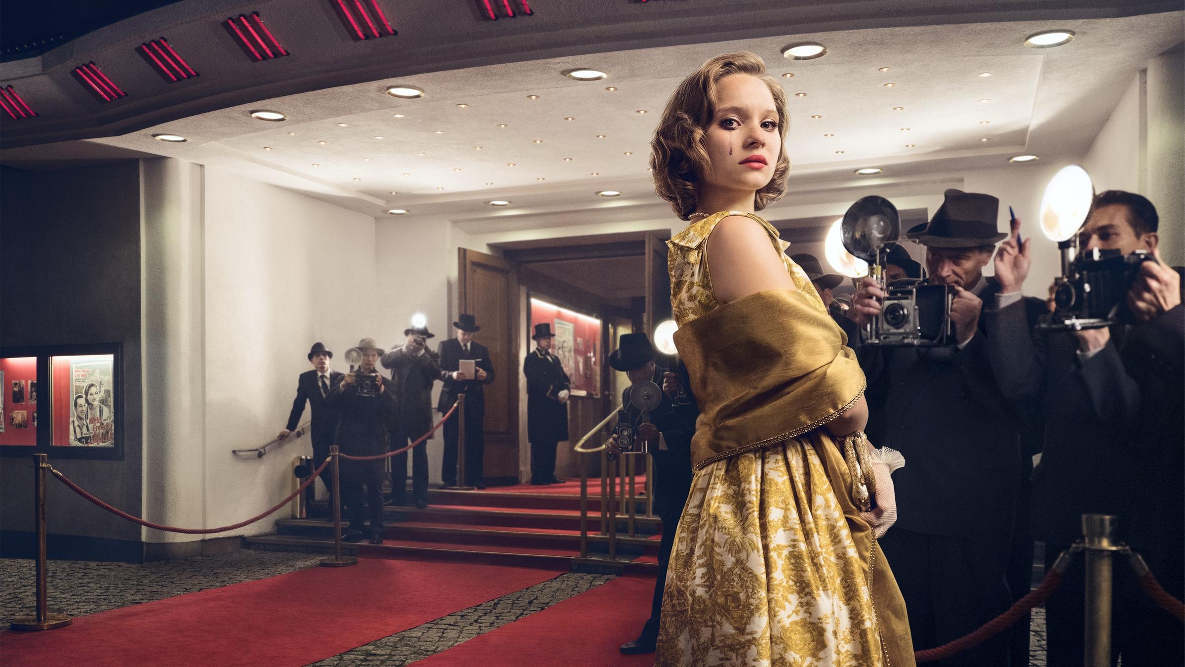 Das Bild zeigt eine Frau in einem Abendkleid und eine Gruppe von Fotografen vor einem Kinoeingang der späten 50er Jahre