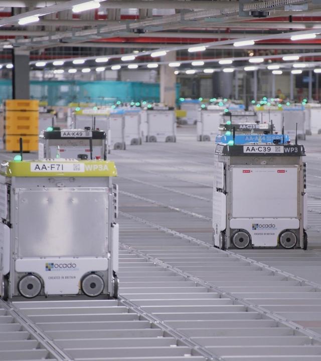"Künstliche Intelligenz - Wettlauf der Großmächte": Dutzende, einem Schrank ähnliche Fahrzeuge mit Blinklichtern, fahren auf Schienen in einer großen mit Neonröhren beleuchteten Halle.