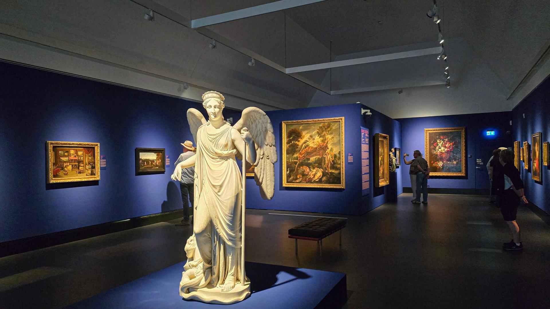 In einem abgedunkelten Ausstellungsraum mit blau gestrichenen Wänden steht in der Mitte eine große Marmorstatue mit Flügeln. An den Wänden hängen Ölgemälde, die ein paar undeutlich erkennbare Besucher betrachten.