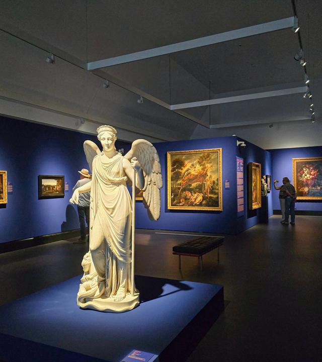 In einem abgedunkelten Ausstellungsraum mit blau gestrichenen Wänden steht in der Mitte eine große Marmorstatue mit Flügeln. An den Wänden hängen Ölgemälde, die ein paar undeutlich erkennbare Besucher betrachten.