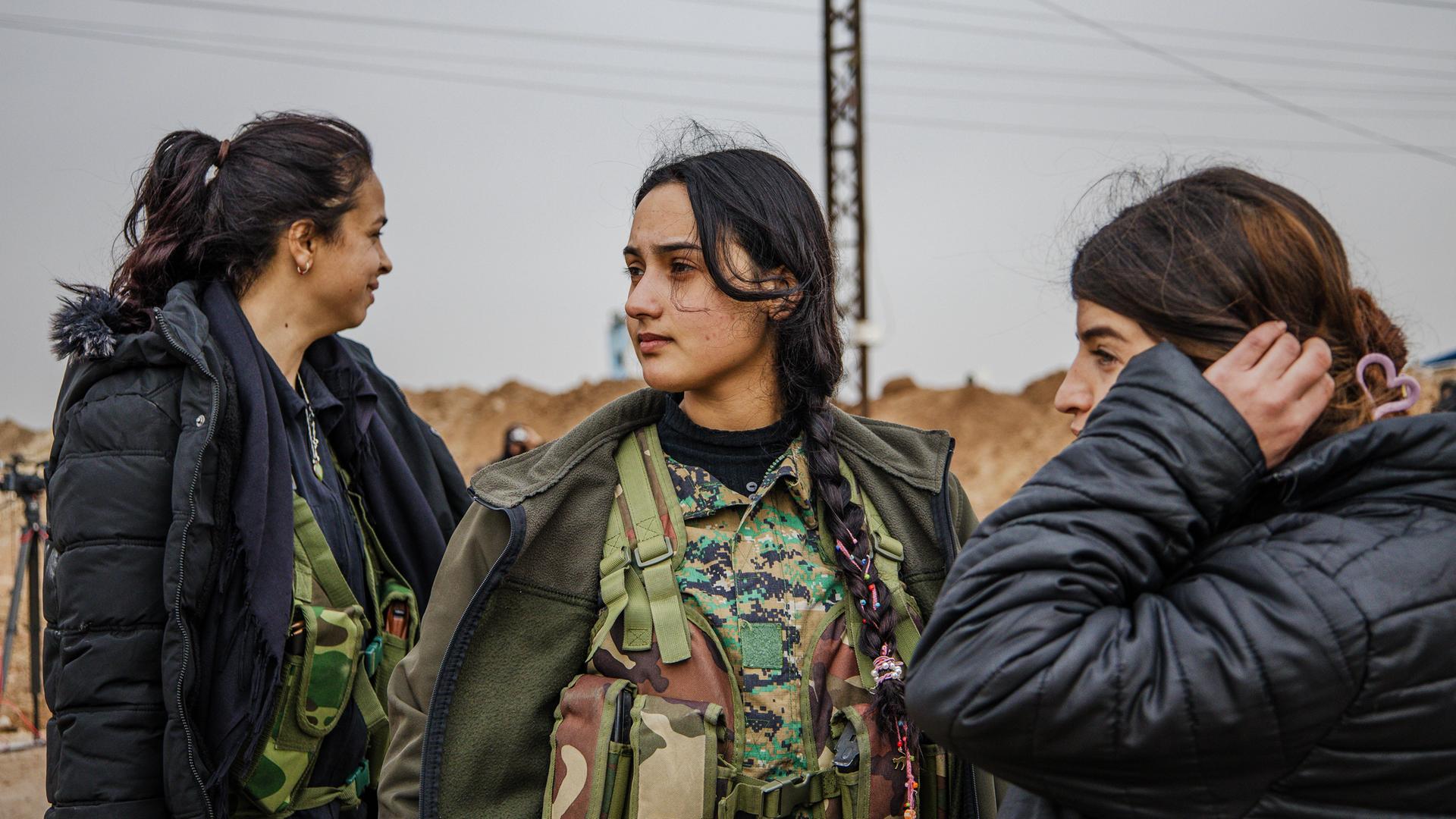 Drei junge Frauen mit dunklen Haaren tragen dicke schwarze Winterjacken über militärischen Camouflage-Westen, stehen nebeneinander und schauen in verschiedene Richtungen.