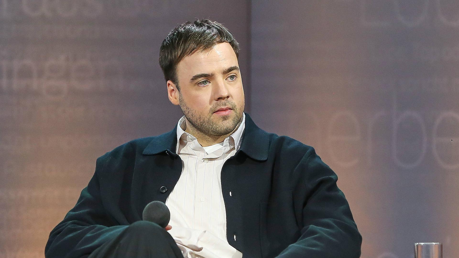 Ein Mann sitzt mit überschlagenen Beinen auf einer Bühne und hält ein Mikrofon in der Hand, während er einer zweiten Person, die nicht im Bild zu sehen ist, zuhört.