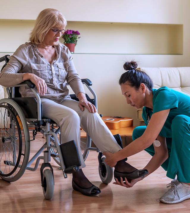 Pflegerin zieht einer Frau im Rollstuhl einen Schuh an