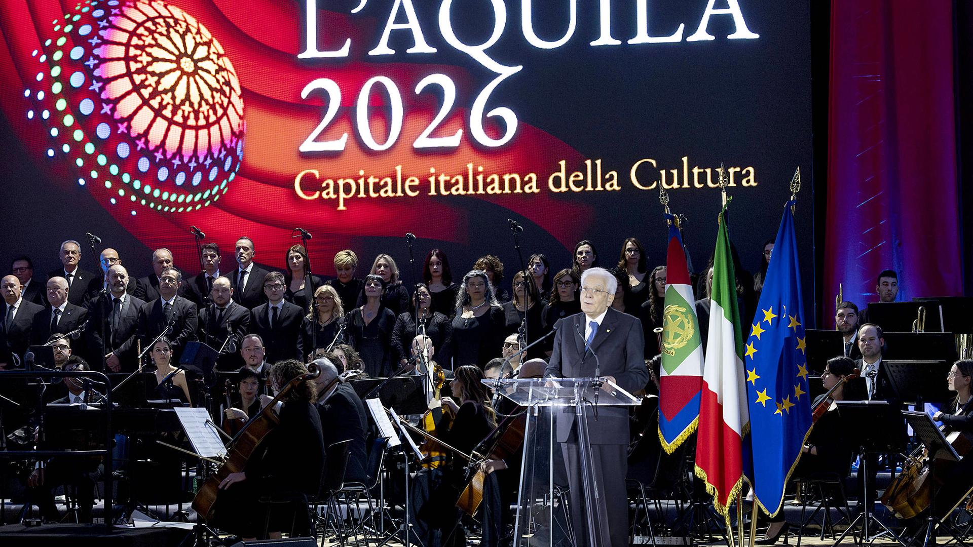 An einem Podest aus Plexiglas neben einem Orchester steht ein Mann und spricht. Im Hintergrund ist groß die Schrift "L'Aquila 2026 Capitale italiana della Cultura auf buntem Hintergrund zu sehen.