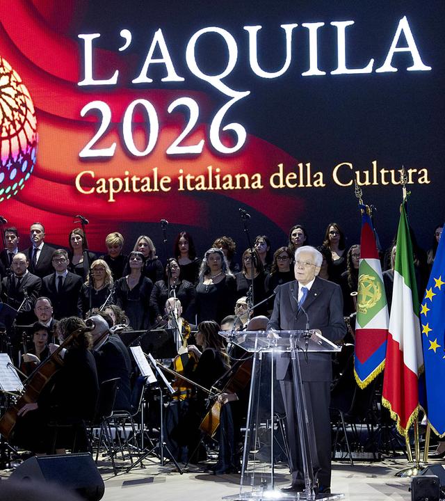 An einem Podest aus Plexiglas neben einem Orchester steht ein Mann und spricht. Im Hintergrund ist groß die Schrift "L'Aquila 2026 Capitale italiana della Cultura auf buntem Hintergrund zu sehen.
