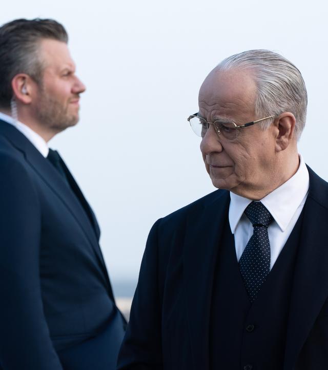 Im Vordergrund steht ein älterer Mann im Anzug mit Brille, der nachdenklich zur Seite schaut. Hinter ihm steht sein Bodyguard, ein Mann mittleren Alters, ebenfalls im Anzug und Knopf im Ohr.