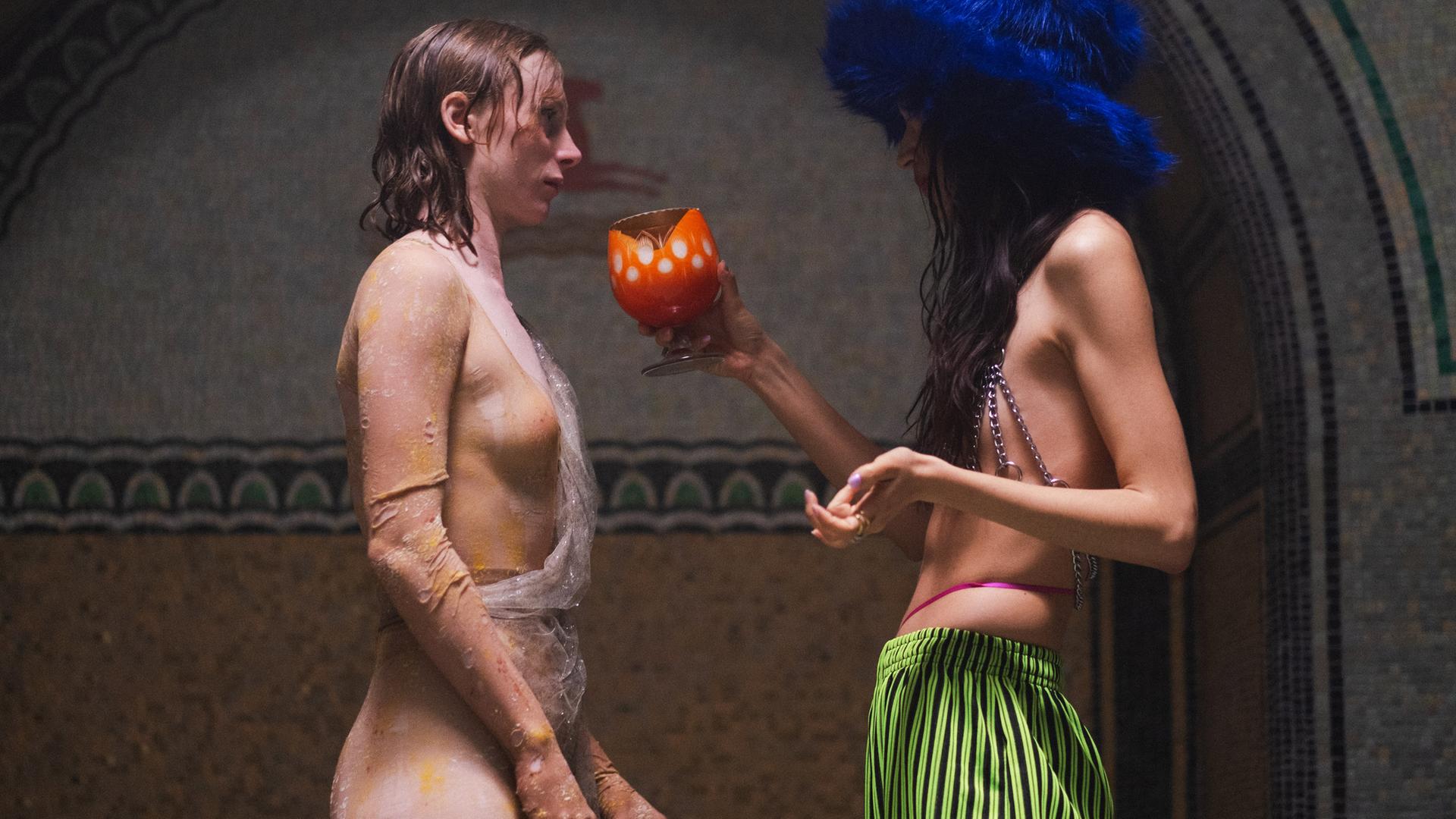 Im Profil sind zwei junge Frauen zu sehen, die sich gegenüber stehen. Eine hat rötliches, nasses Haar und trägt eine Art durchsichtige Schlangenhaut, die andere trägt eine auffällige grüne Hose, ein Kettenoberteil und einen blauen Plüschhut, ihr Haare ist lang und dunkel, ihr Gesicht sieht man nicht. Sie hält der änderen einen orangefarbenen Kelch hin.
