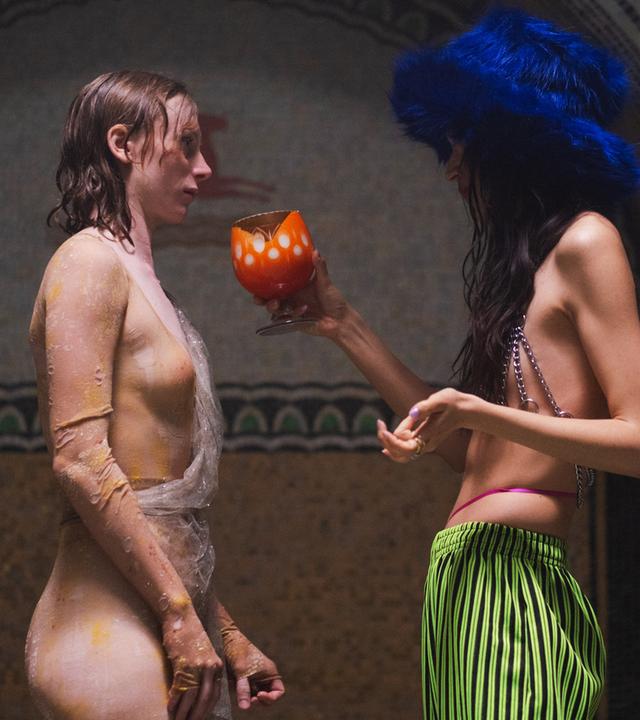 Im Profil sind zwei junge Frauen zu sehen, die sich gegenüber stehen. Eine hat rötliches, nasses Haar und trägt eine Art durchsichtige Schlangenhaut, die andere trägt eine auffällige grüne Hose, ein Kettenoberteil und einen blauen Plüschhut, ihr Haare ist lang und dunkel, ihr Gesicht sieht man nicht. Sie hält der änderen einen orangefarbenen Kelch hin.