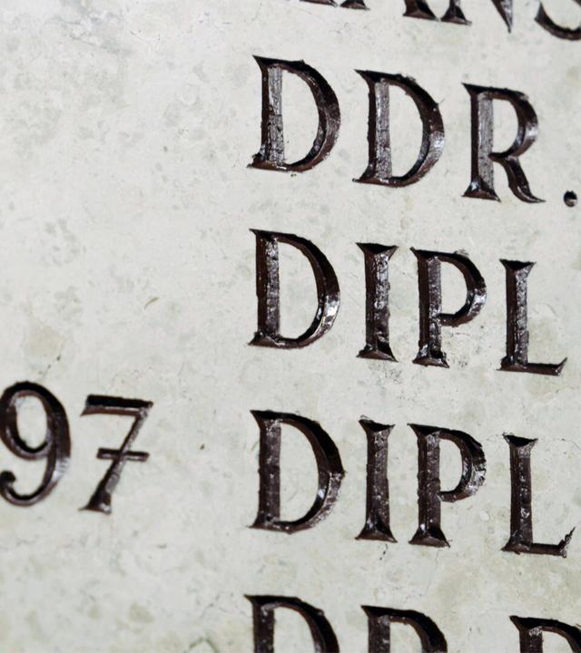 Das Bild zeigt einen Ausschnitt einer Gedenktafel oder Plakette aus einem hellen Material, das möglicherweise in Stein gemeißelt ist. Die Oberfläche der Tafel ist texturiert und zeigt eine Vielzahl von eingravierten Buchstaben und Zahlen.   Die Inschrift enthält akademische Titel, darunter "DDR. DR.", "DIPL. ING." und "DIPL. KFM.", sowie die Jahreszahl "1997". Die Buchstaben sind in Großbuchstaben und zeichnen sich durch eine kräftige, dreidimensionale Form aus, wodurch sie gut lesbar sind. Die Buchstaben scheinen dunkel eingelassen zu sein, was einen starken Kontrast zur hellen Oberfläche schafft.   Die umgebende Fläche der Tafel wirkt rau und hat einige kleine Risse oder Abnutzungen, die auf ein gewisses Alter hindeuten. Das Gesamtbild vermittelt den Eindruck von formeller Ehrung und akademischer Traditionspflege.