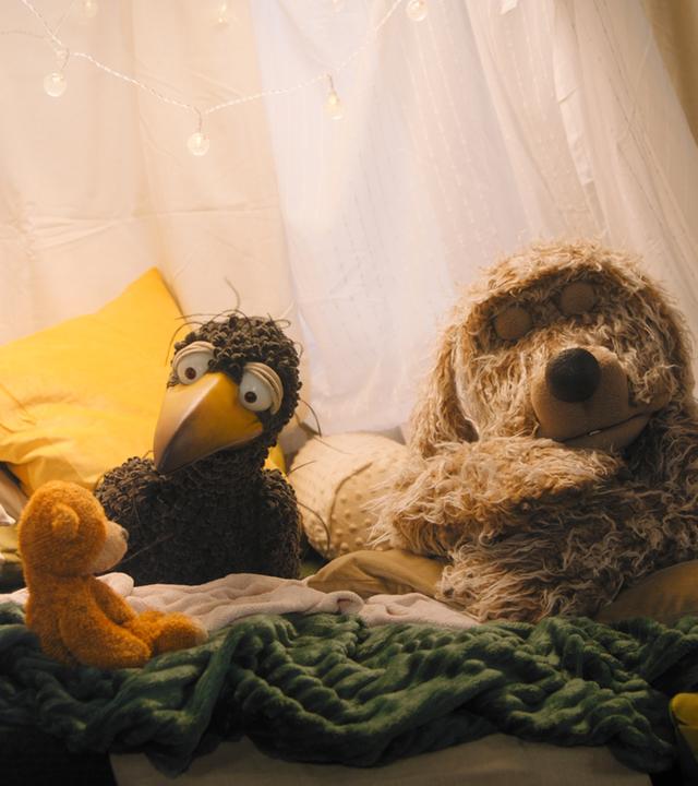 Rabe Rudi hockt in der gemütlichen Kuschelecke. Er schaut einen orangefarbenen Teddybären an. Rechts daneben schläft Hund Buffo.