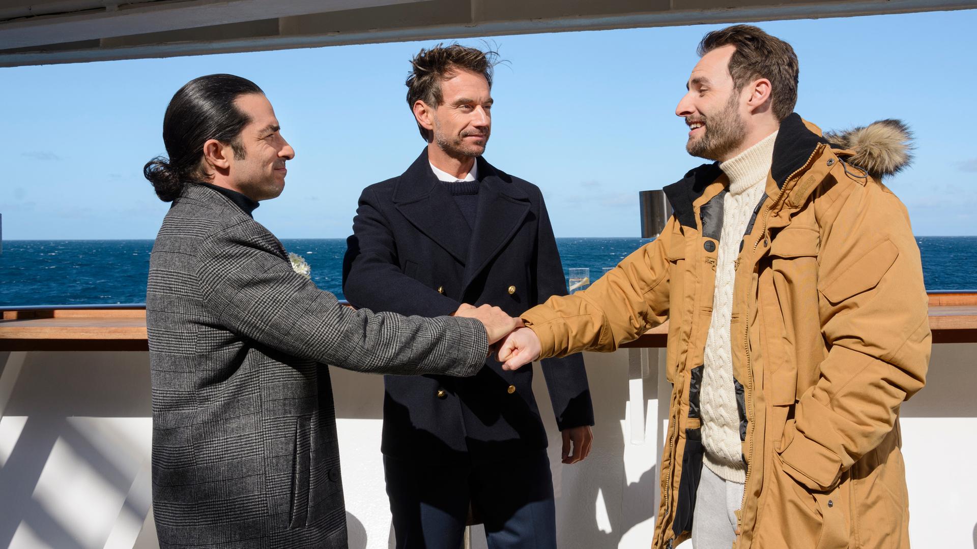"Das Traumschiff: Lappland": An Deck des Schiffs stehen Max Parger (Florian Silbereisen), Patrick Steiner (Daniel Fritz) und Maik Berghammer (Tayfun Baydar) zusammen. Die Freunde legen die Hände übereinander und lächeln sich an.
