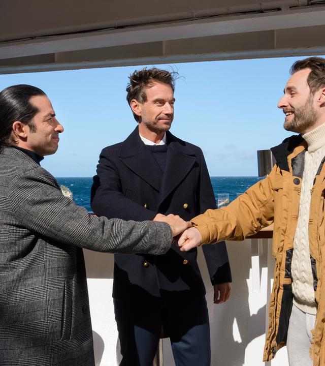 "Das Traumschiff: Lappland": An Deck des Schiffs stehen Max Parger (Florian Silbereisen), Patrick Steiner (Daniel Fritz) und Maik Berghammer (Tayfun Baydar) zusammen. Die Freunde legen die Hände übereinander und lächeln sich an.
