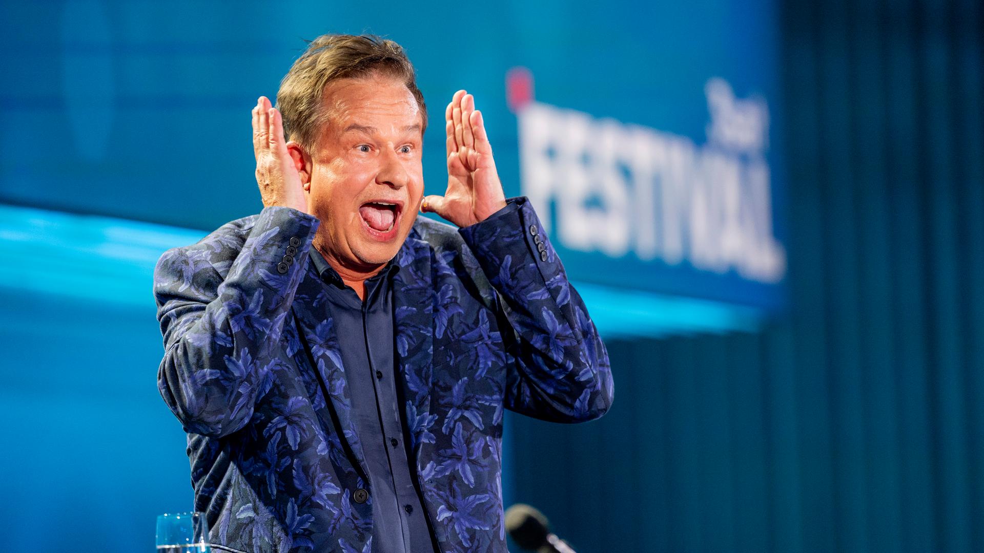 Kabarettist steht auf der linken Bildseite. Er hebt beide Hände neben dem Kopf. Sein Mund ist offen. Er trägt einen blauen Anzug mit Muster und darunter ein dunkelblaues Hemd. Die Bühne ist blau beleuchtet.