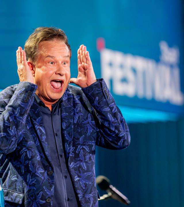 Kabarettist steht auf der linken Bildseite. Er hebt beide Hände neben dem Kopf. Sein Mund ist offen. Er trägt einen blauen Anzug mit Muster und darunter ein dunkelblaues Hemd. Die Bühne ist blau beleuchtet.