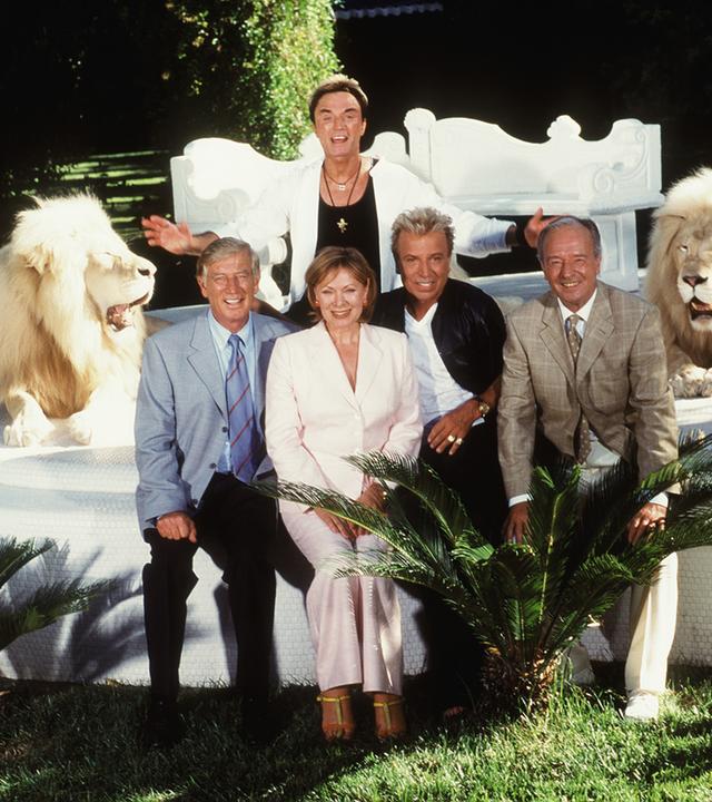 "Das Traumschiff - Las Vegas": Kapitän Paulsen (Siegfried Rauch), Horst Schröder (Horst Naumann) und Beatrice (Heide Keller) sitzen mit Siegfried (Siegfried Fischbacher) und Roy (Roy Horn) eingerahmt von zwei Löwen auf einer Steinplattform.
