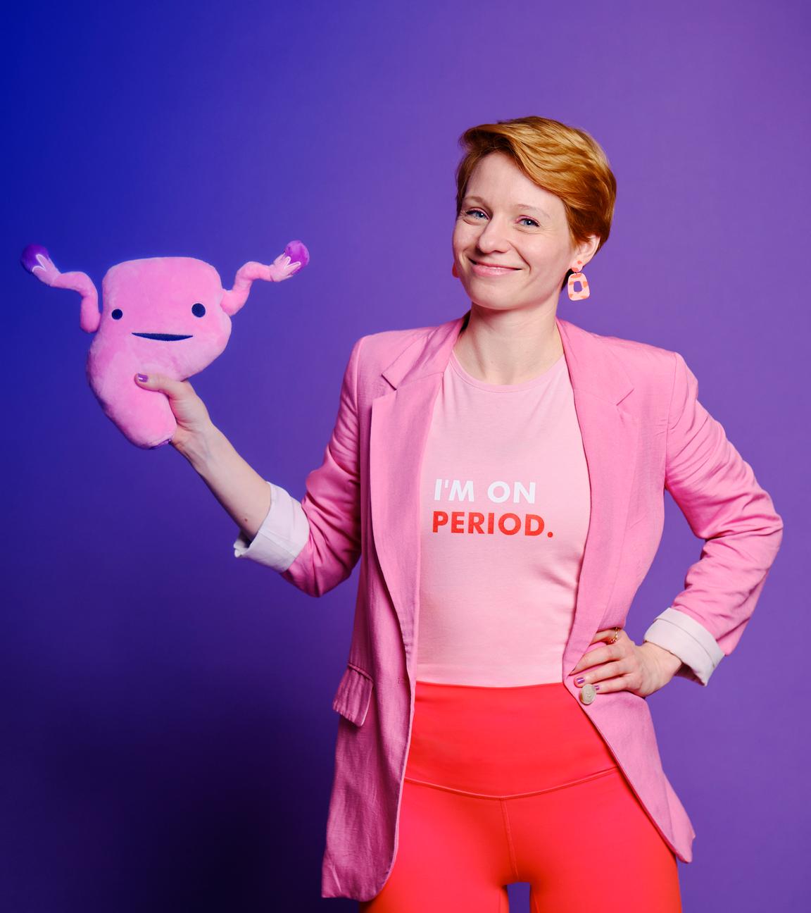 Steffi Platt steht vor einer lilablauen Wand. Sie hat eine orangefarbene Leggings und einen rosa Blazer an. Außerdem trägt sie ein T-Shirt mit der Aufschrift: "I‘m on period.". In der rechten Hand hält sie eine Gebärmutter aus Plüsch. Ihren linken Arm hat sie in die Hüfte gestemmt. Sie lächelt in die Kamera.