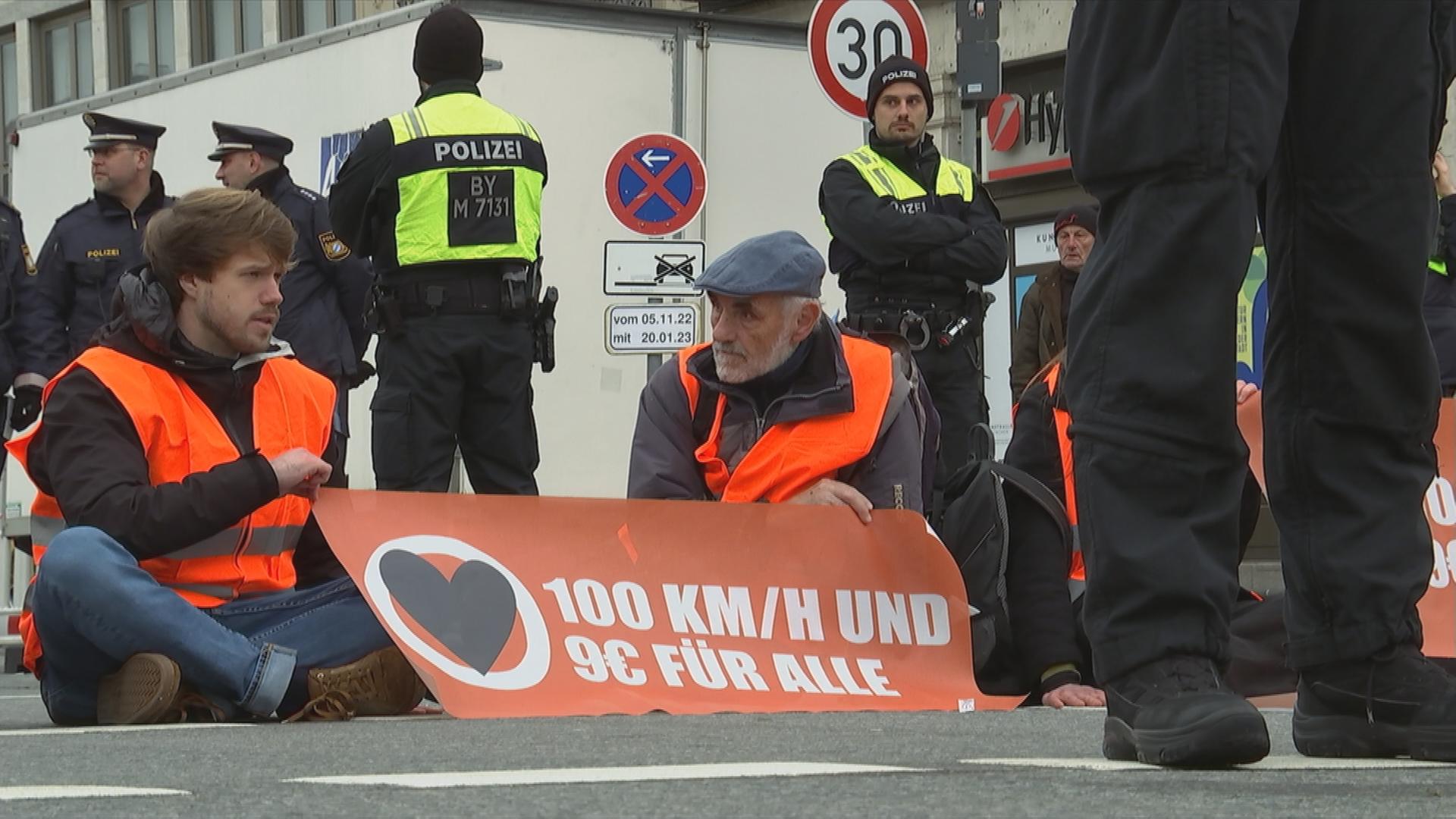 Zwei Männer sitzen in orange farbenen Warnwesten auf dem Asphalt und halten ein Plakat hoch. Sie sind umzingelt von Polizei.