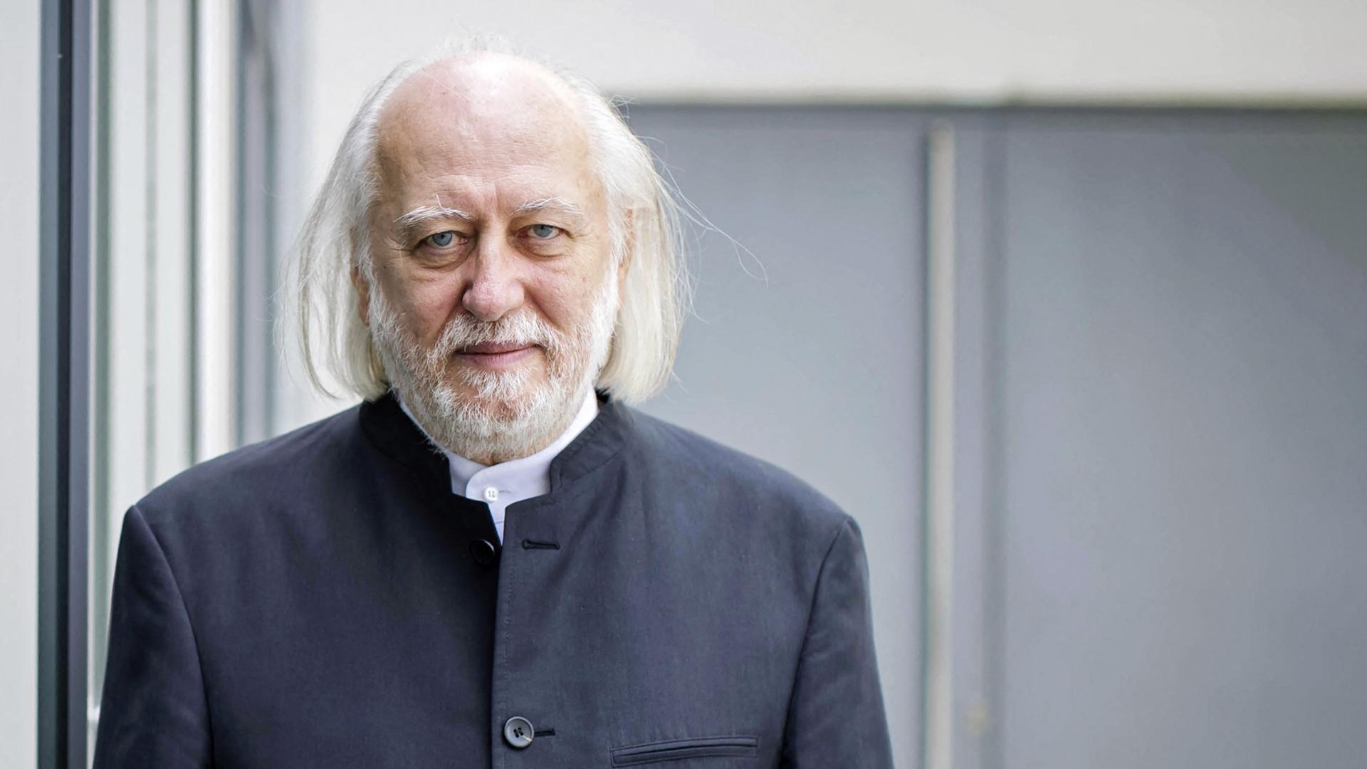 Porträtfoto eines älteren Mannes mit Glatze und schütterem schulterlangen weißen Haaren und graumeliertem Bart blickt lächelnd in die Kamera.