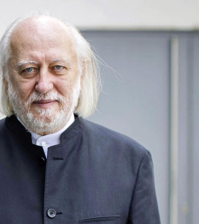 Porträtfoto eines älteren Mannes mit Glatze und schütterem schulterlangen weißen Haaren und graumeliertem Bart blickt lächelnd in die Kamera.