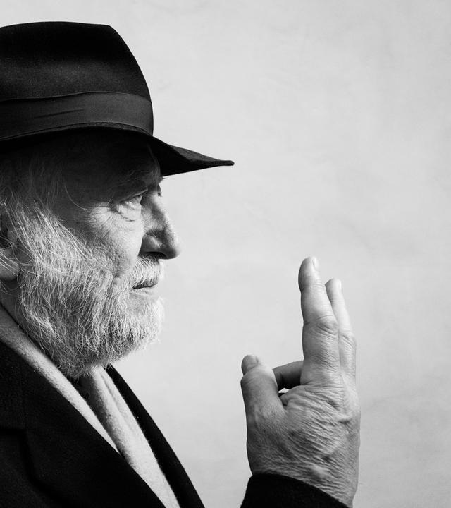 Schwarzweißes Profilfoto eines älteren Mannes mit grauen Haaren und Bart, der einen schwarzen Hut trägt und die rechte Hand mit zu einem V gespreizten Zeige- und Mittelfinger vor sich hält.