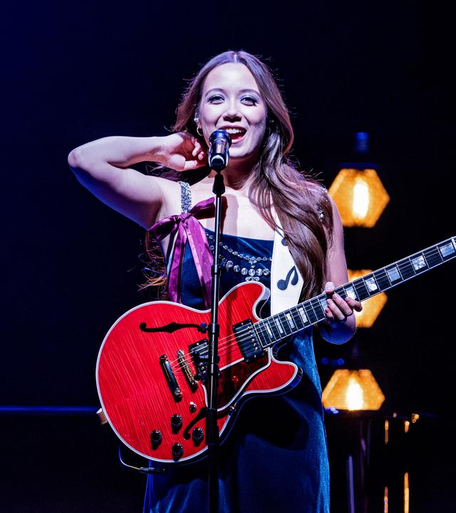 Singer-Songwriterin Laufey steht vor einem Mikrofon, hat eine Gitarre in der Hand und lächelt.