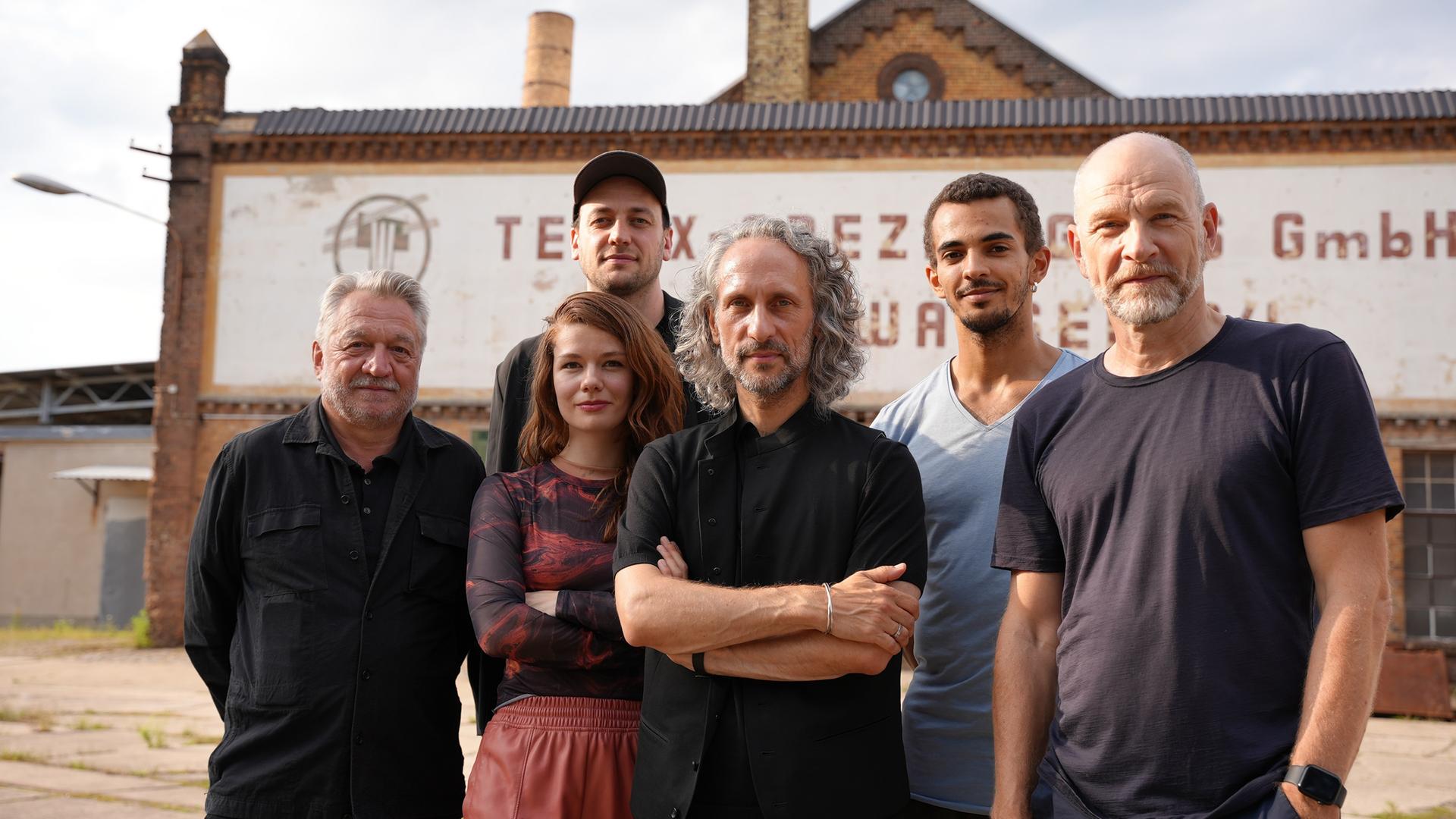 Sechs Personen stehen vor einem alten, industriell anmutenden Gebäude. Der Mann links trägt ein schwarzes Hemd und hat graues Haar. Neben ihm steht eine Frau mit langen, rötlichen Haaren, die ein gemustertes Oberteil und eine rotbraune Hose trägt. Daneben ist ein Mann mit mittellangem, grauem, lockigem Haar und schwarzer Kleidung, der die Arme verschränkt hat zu sehen. In der hinteren Reihe stehen von links nach rechts ein Mann mit schwarzer Kappe und schwarzem Hemd, ein jüngerer Mann mit kurzem, dunklem Haar in einem blauen T-Shirt und ein älterer Mann mit kahlrasiertem Kopf und einem dunkelblauen T-Shirt