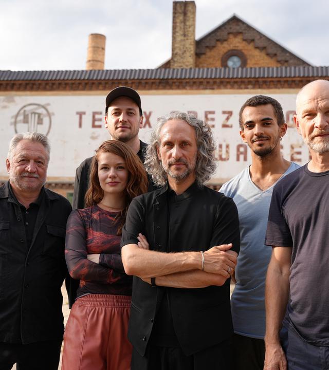 Sechs Personen stehen vor einem alten, industriell anmutenden Gebäude. Der Mann links trägt ein schwarzes Hemd und hat graues Haar. Neben ihm steht eine Frau mit langen, rötlichen Haaren, die ein gemustertes Oberteil und eine rotbraune Hose trägt. Daneben ist ein Mann mit mittellangem, grauem, lockigem Haar und schwarzer Kleidung, der die Arme verschränkt hat zu sehen. In der hinteren Reihe stehen von links nach rechts ein Mann mit schwarzer Kappe und schwarzem Hemd, ein jüngerer Mann mit kurzem, dunklem Haar in einem blauen T-Shirt und ein älterer Mann mit kahlrasiertem Kopf und einem dunkelblauen T-Shirt