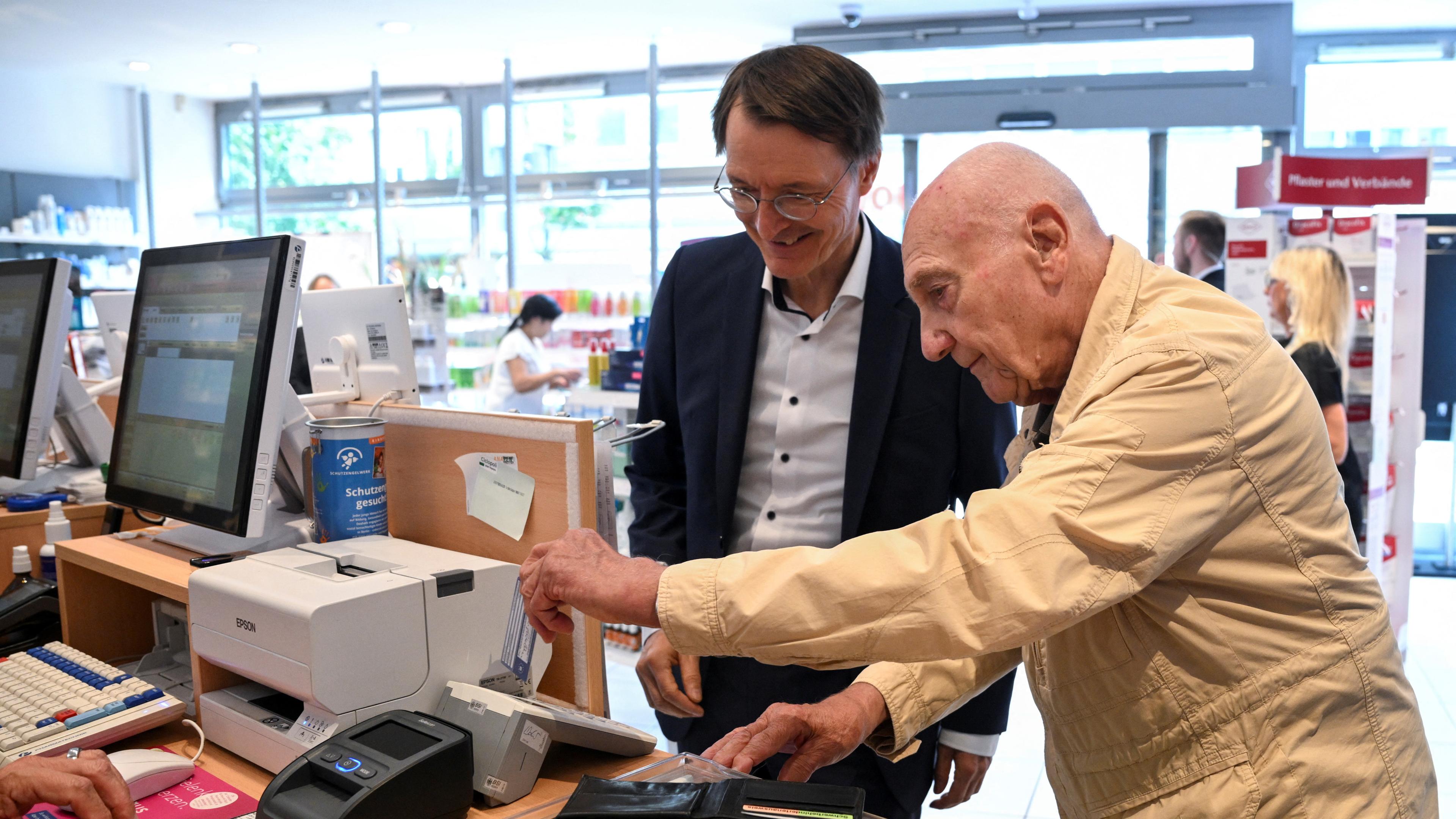 Karl Lauterbach neben einem alten Mann, derin einer Apotheke eine Karte in ein Kartenlesegerät steckt