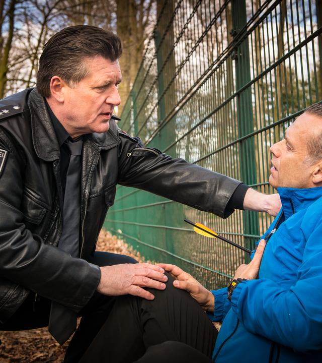 "Notruf Hafenkante - Lautlos": Der Polizeibeamte Hans Moor (Bruno F. Apitz) kniet vor einem mit einem Pfeil angeschossenen Mann, der auf dem Waldboden sitzt und mit dem Rücken an einen Zaun lehnt. Er trägt eine Sportjacke.