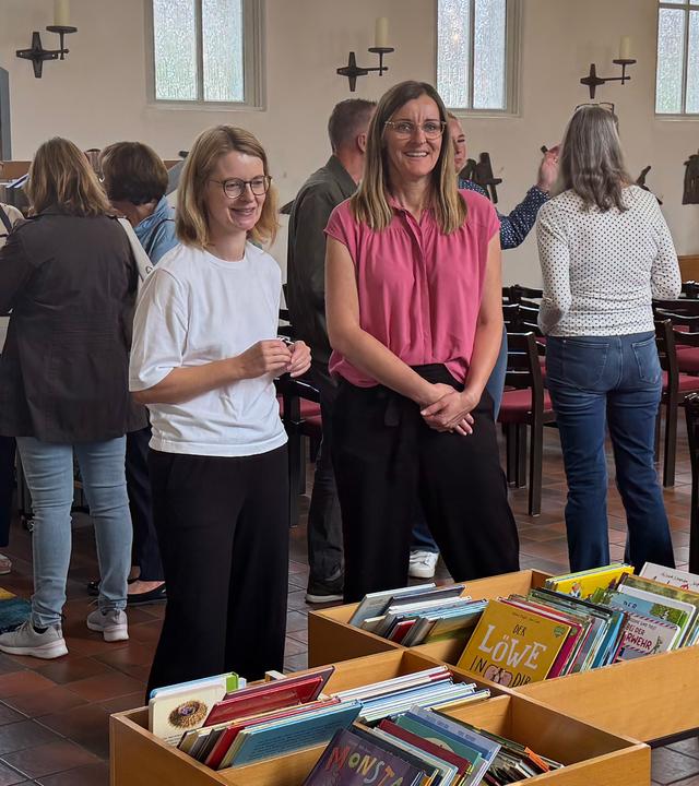 Mehrere Menschen stehen und sprechen in der zur Bücherei umgestalteten Kirche miteinander. Im Vordergrund stehen Regale und Kisten voller Kinderbücher, daneben stehen Stefanie Hayen und Stefanie Eilers vom Team von "Gemeinsam statt einsam".