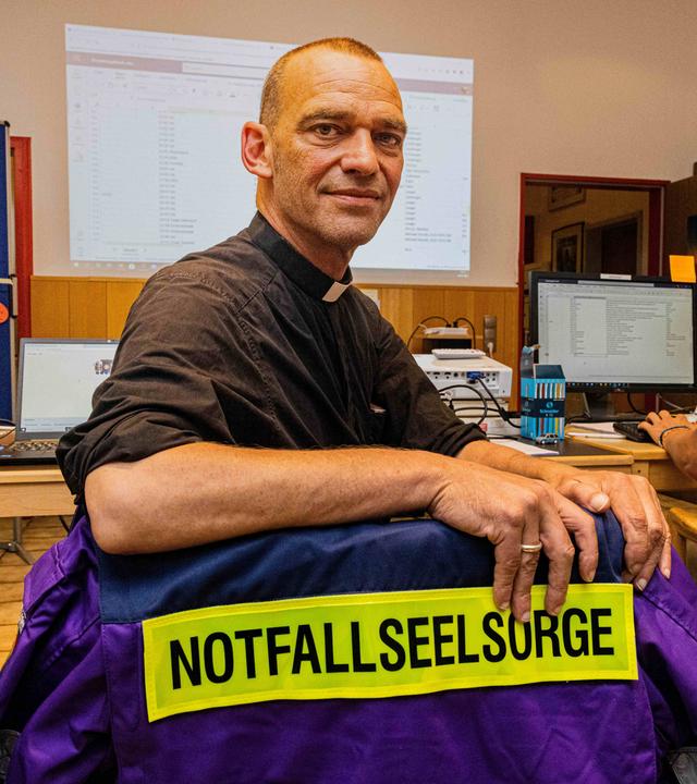 Albrecht Roebke sitzt in einem Büro an einem Tisch. Über seinen Stuhl hängt eine lilafarbene Einsatzjacke mit der Aufschrift "Notfallseelsorge". Er blickt über die Schulter in die Kamera. Neben ihm arbeitet eine weitere Person am Computer.