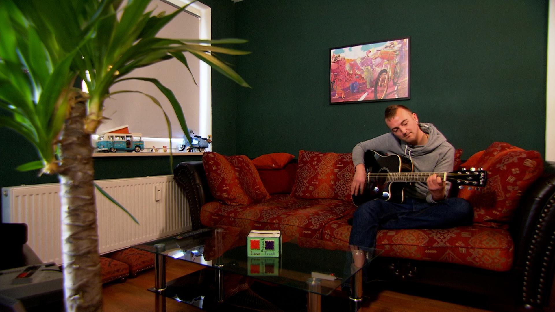 Ein junger Mann sitzt auf einem roten Sofa und spielt Gitarre.