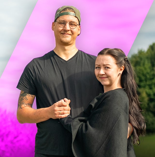 Auf der rechten Bildseite steht ein Mann und eine Frau eng beieinander, sie halten sich an der Hand. Auf der linken Bildseite ist ein zerstörtes Auto am Straßenrand nach einem Unfall zu sehen.