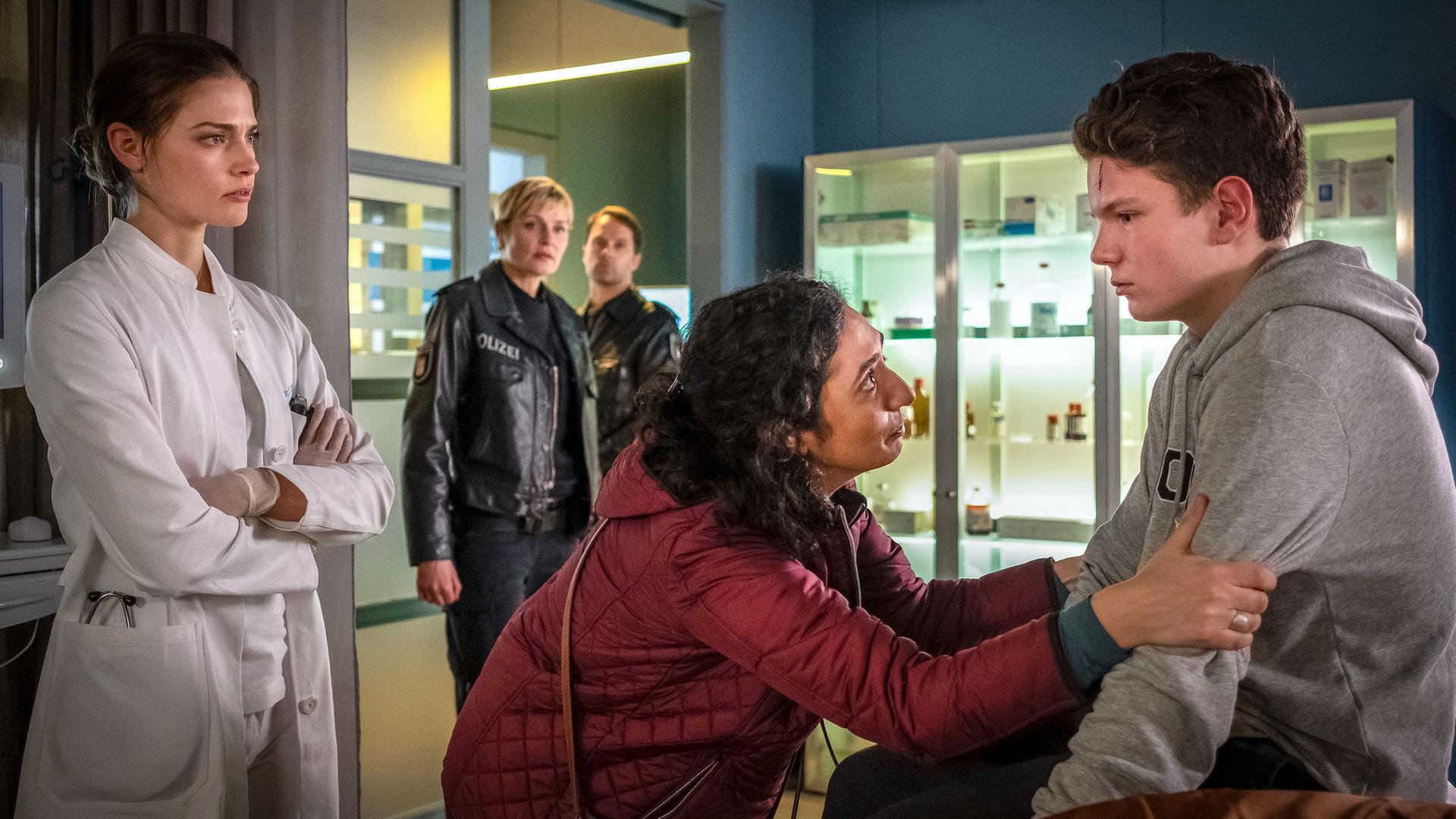 "Notruf Hafenkante - Lebend wieder raus": Emil Grönhoff (Julius Störmer) sitzt in einem Behandlungsraum des EKH auf einer Behandlungsliege. Vor ihm steht seine Mutter (Ariella Hirshfeld). Sie hält ihn an seinen Armen fest und sieht ihn flehend an. Hinter ihr steht Dr. Eva Grünberg (Laura Berlin). Sie hat die Arme vorm Oberkörper verschränkt und schaut den Jungen besorgt an. Im Hintergrund stehen die Polizisten Melanie Hansen (Sanna Englund) und Mattes Seeler (Matthias Schloo) und beobachten die Szene.