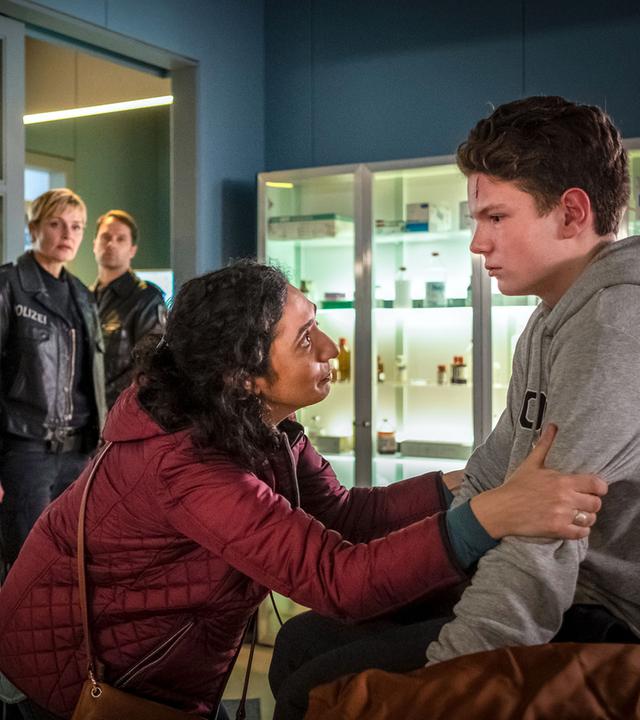 "Notruf Hafenkante - Lebend wieder raus": Emil Grönhoff (Julius Störmer) sitzt in einem Behandlungsraum des EKH auf einer Behandlungsliege. Vor ihm steht seine Mutter (Ariella Hirshfeld). Sie hält ihn an seinen Armen fest und sieht ihn flehend an. Hinter ihr steht Dr. Eva Grünberg (Laura Berlin). Sie hat die Arme vorm Oberkörper verschränkt und schaut den Jungen besorgt an. Im Hintergrund stehen die Polizisten Melanie Hansen (Sanna Englund) und Mattes Seeler (Matthias Schloo) und beobachten die Szene.