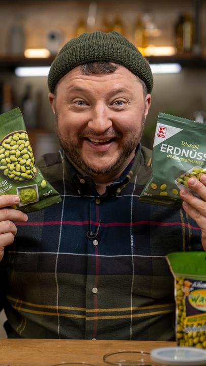 "Lebensmitteltricks - Lege packt aus: Miese Maschen im Snack-Regal": Sebastian Lege hält lachend zwei Wasabi-Nuss-Packungen hoch.