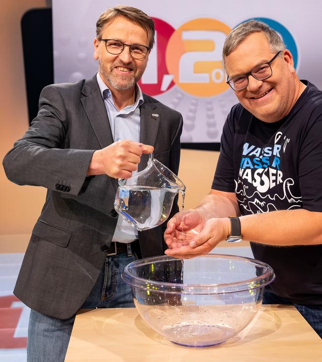 Ökologe Dr. Martin Zimmer steht mit Elton an einem hüfthohen Tisch. Martin Zimmer lässt Elton aus einer Karaffe Wasser über die Hände laufen. Auf dem Tisch steht eine große Schüssel, in dem das Wasser aufgefangen wird.