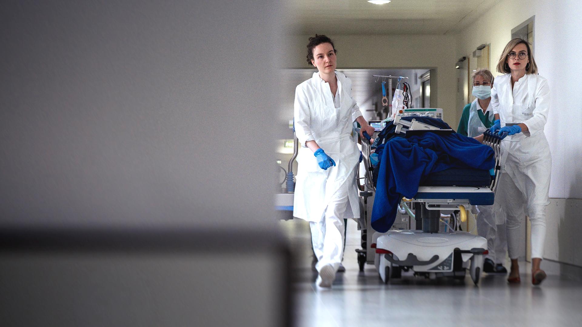 In der linken Bildhälfte ist unscharf eine Säule zu sehen, in der rechten sieht man drei weibliche Notpflegekräfte, die unter Stress im Krankenhaus eine Patientenliege rollen. 