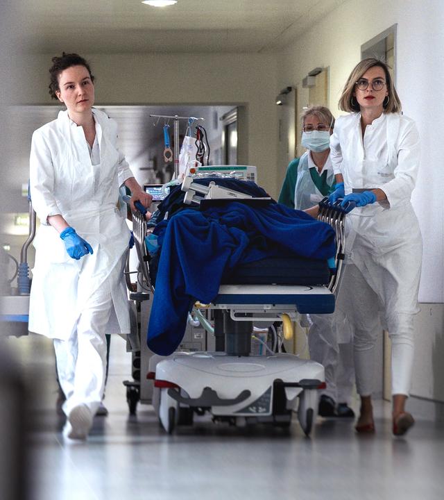 In der linken Bildhälfte ist unscharf eine Säule zu sehen, in der rechten sieht man drei weibliche Notpflegekräfte, die unter Stress im Krankenhaus eine Patientenliege rollen. 