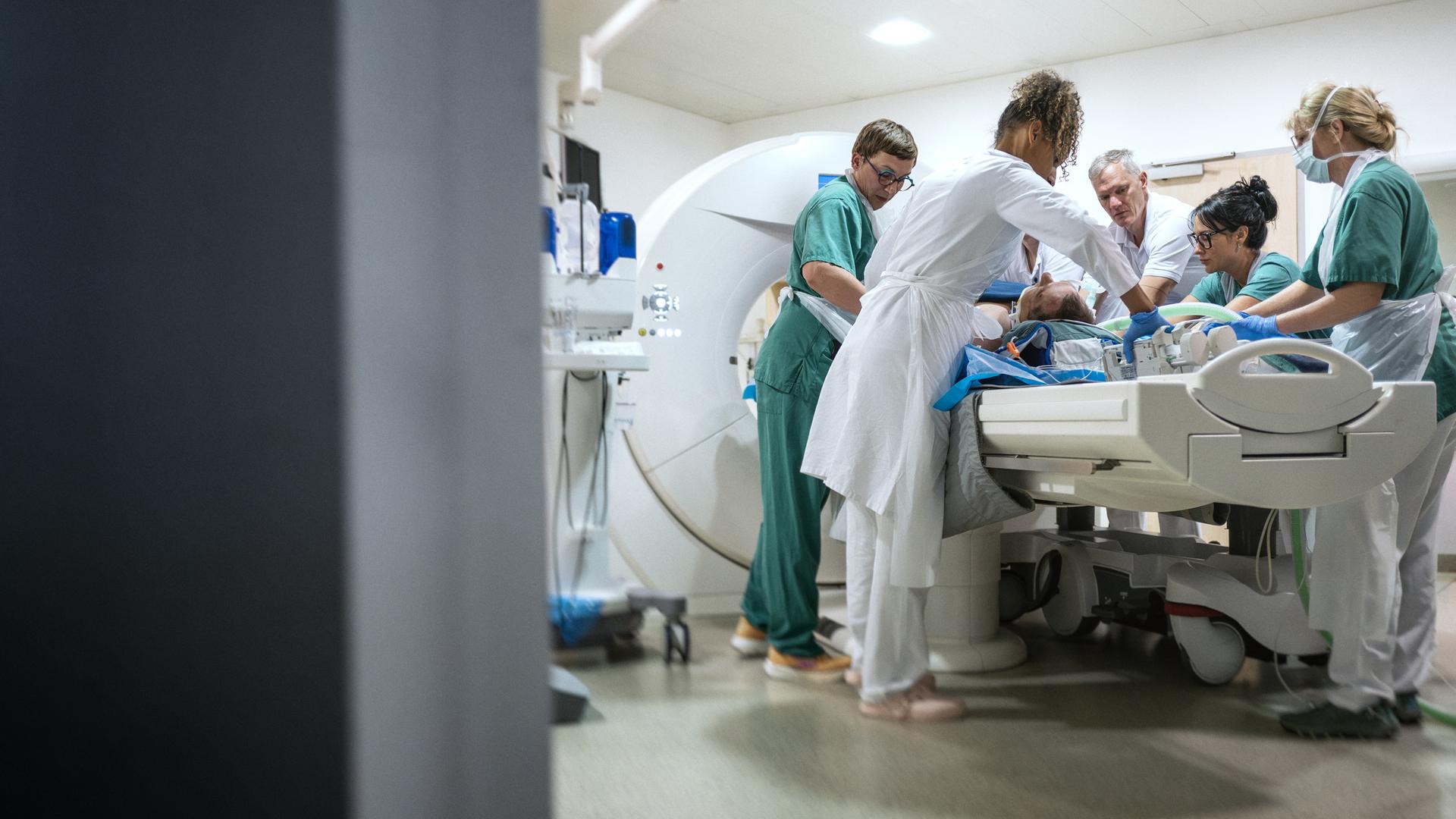 Am linken Bildrand ist der Türrahmen unscharf zu sehen. Wir blicken in einen Raum mit MRT-Röhre, wo fünf Sanitäter und Ärzte einen Patienten für die Untersuchung präparieren.