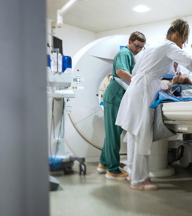 Am linken Bildrand ist der Türrahmen unscharf zu sehen. Wir blicken in einen Raum mit MRT-Röhre, wo fünf Sanitäter und Ärzte einen Patienten für die Untersuchung präparieren.