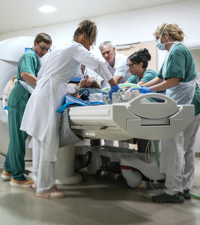 Am linken Bildrand ist der Türrahmen unscharf zu sehen. Wir blicken in einen Raum mit MRT-Röhre, wo fünf Sanitäter und Ärzte einen Patienten für die Untersuchung präparieren.